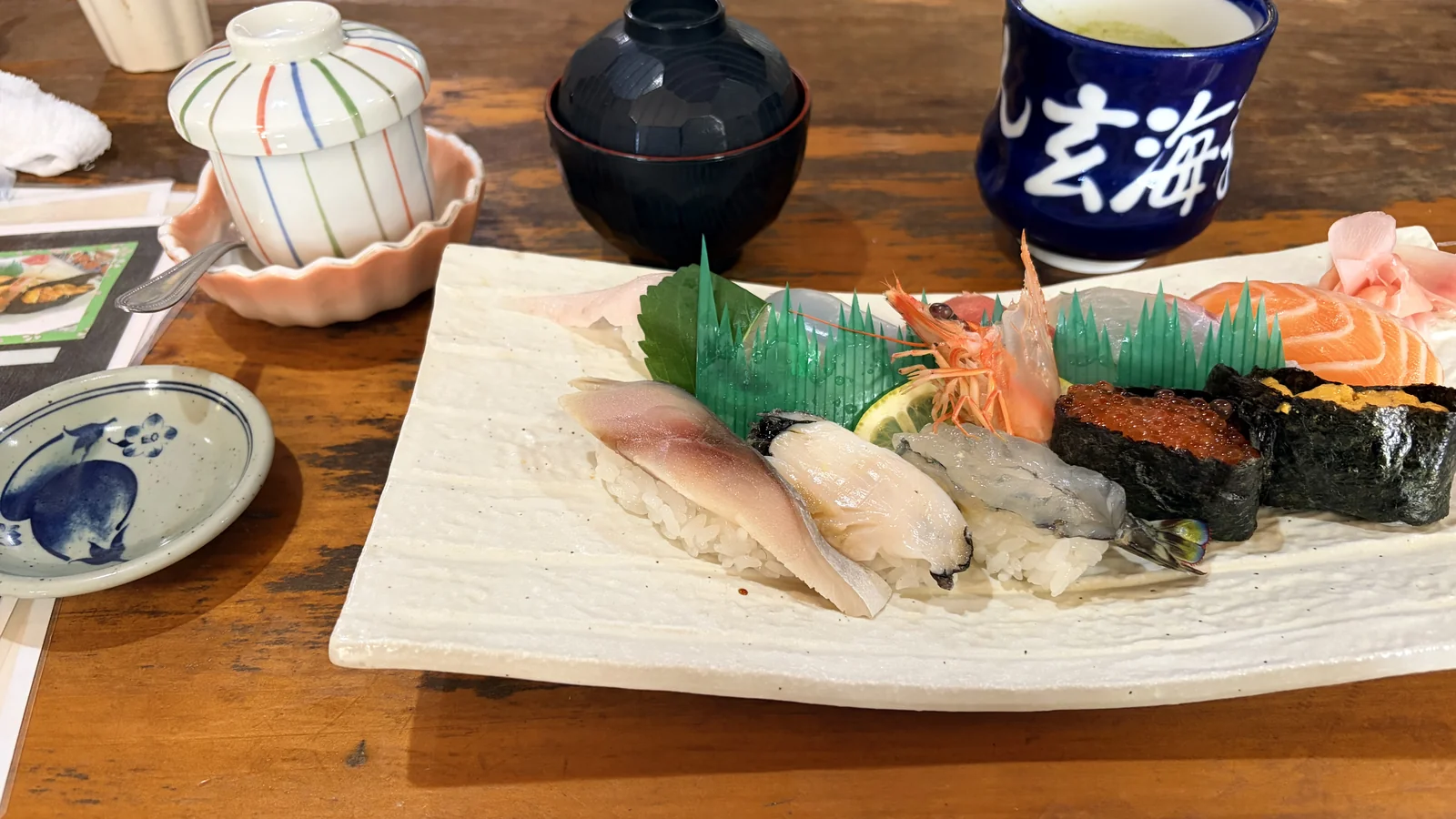 ひぜん玄海寿しのおまかせ握り。白い角皿に光り物、白身、剥き身のエビ、いくらとうにの軍艦、サーモンが並び、エビの後ろに頭が飾りとして立てられている