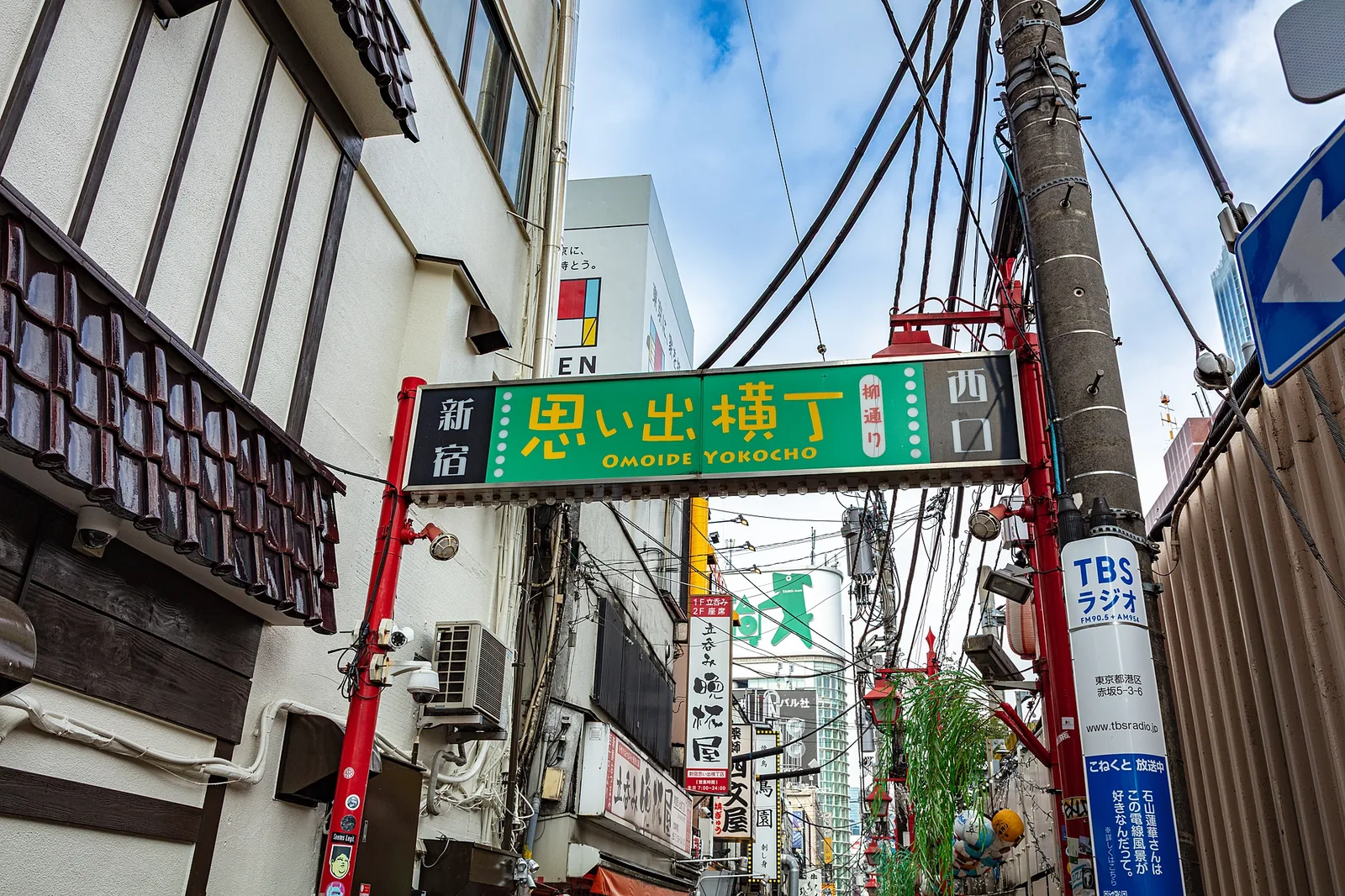 新宿西口・思い出横丁の入口。赤い柱に支えられた緑色の看板に「新宿 思い出横丁 西口」「OMOIDE YOKOCHO」の文字。頭上には電線が張り巡らされている